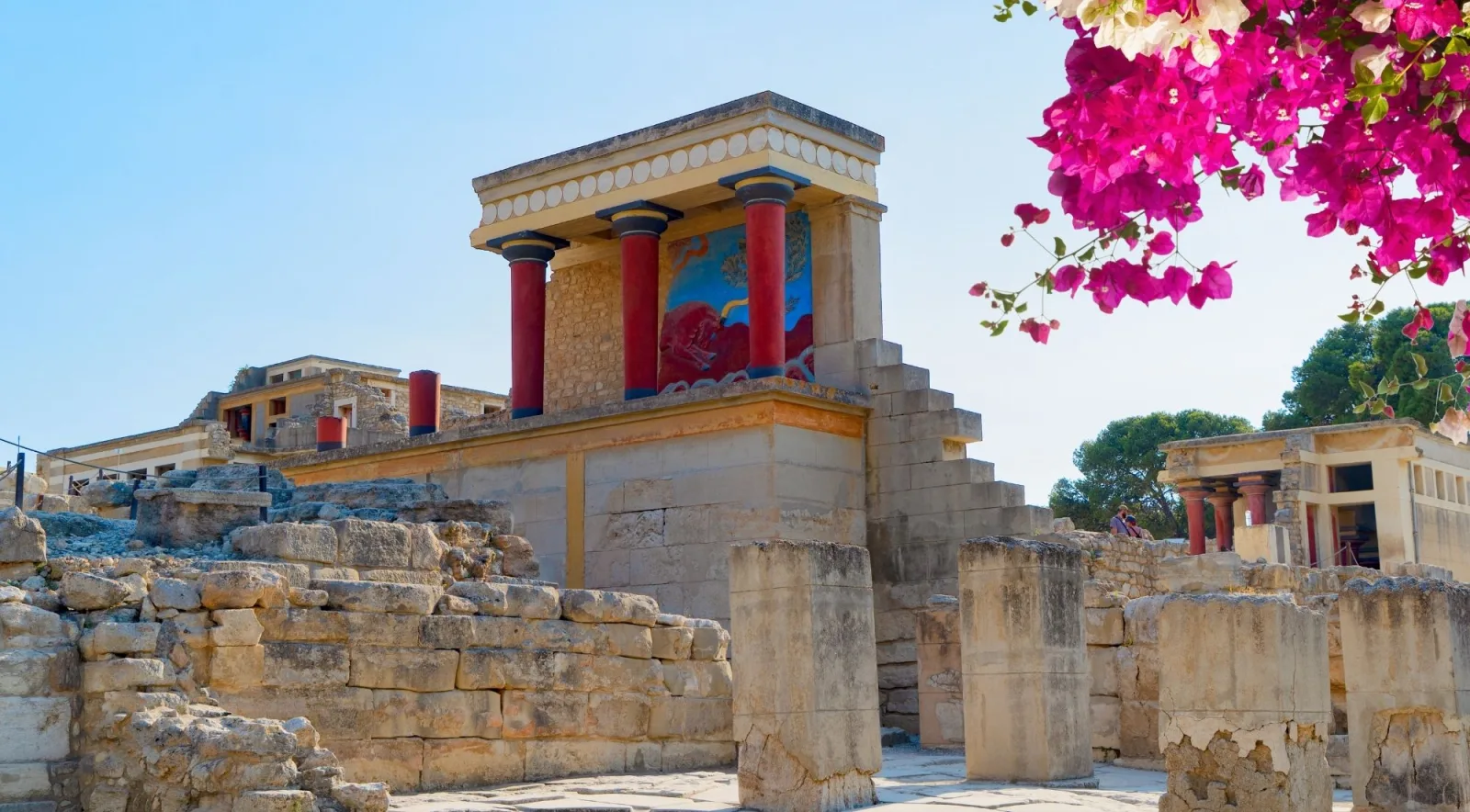 The restored North Entrance bastion at Knossos, with red Minoan columns and the charging-bull fresco, framed by bougainvillea