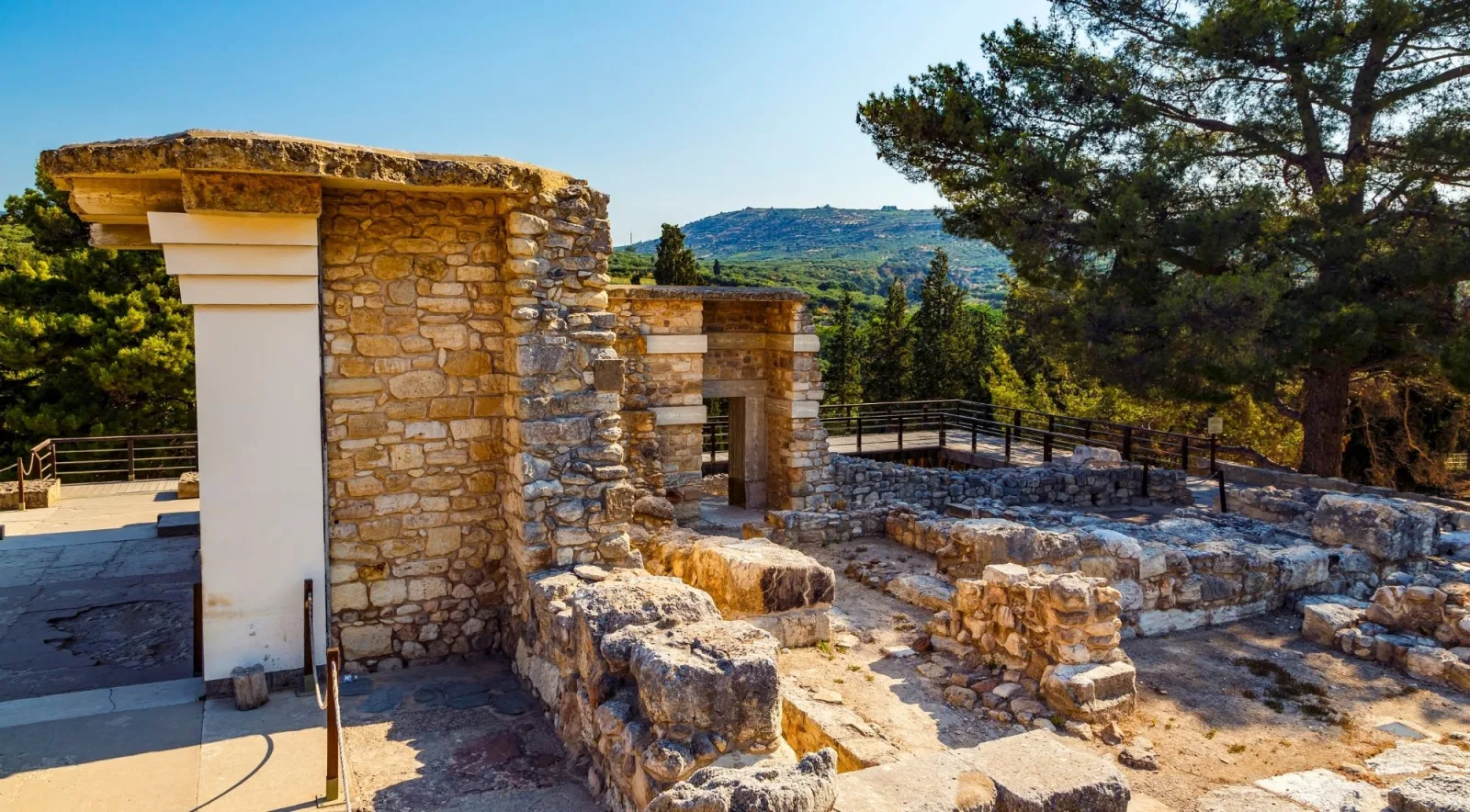 Restored doorways and stone walls at the edge of the Knossos site, with pine trees and hills beyond
